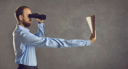 Profile view of serious young guy who needs glasses looking in binoculars and reading book hes...