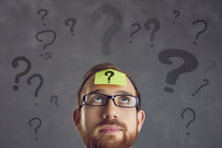 A question mark on a piece of paper on the forehead of a businessman with glasses. A young man thinks about a decision. Young guy in glasses with yellow post it sticky note on forehead looking up at empty advertising copy spaceの写真素材