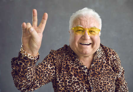Close up of cheerful stylish glamorous senior man showing v-sign peace symbol on gray background....
