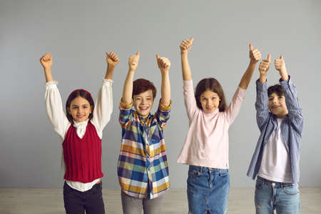 Energetic optimistic preteen children happily smiling show hands up with thumbs up gesture. Victory, confidence, success and approval concept. Studio portrait shot on grey copy spaceの写真素材