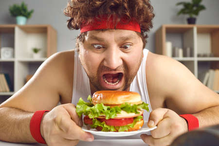 Hungry crazy chubby man in sportswear sitting at table and eating fast food hamburger. Overweight male person junk meal lover with opened mouth widely closeup portrait. Diet failure, cheat-mealの写真素材