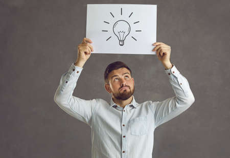 Handsome man holding a sheet of paper with a picture of an Edison light bulb. Young guy thinking and considering an interesting innovative idea he could use for his businessの写真素材