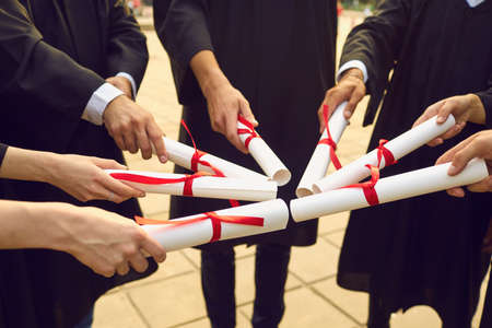 Hands of university graduates in traditional black masters mantles holding dimplomas with honors in sun shape and celebrating finishing universityの写真素材