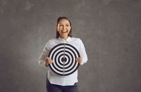Young loudly laughing emotional caucasian woman holding dart board target standing on studio wall and looking aside. Business startup or project success, strategy planning, targeting marketing conceptの写真素材