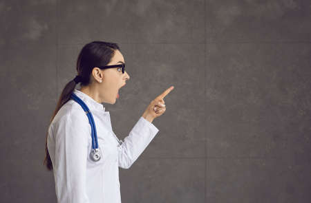 Shocked young caucasian doctor wearing uniform and stethoscope standing with open mouth amazed facial expression surprised pointing with hand finger forward view studio shot portraitの写真素材