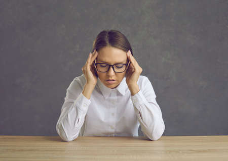 Tired young caucasian woman office worker, student or teacher suffering from headache sitting at desk over studio background. Upset unhappy pretty charming freelancer feeling bad touching templeの写真素材