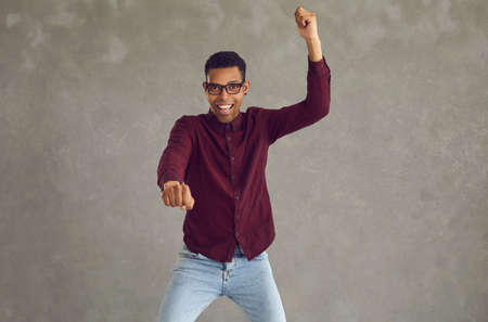Young african american man rejoicing success with clenched fists moving like riding horse of bike studio shot. Black happy and excited male student showing winner expression celebrating victoryの写真素材