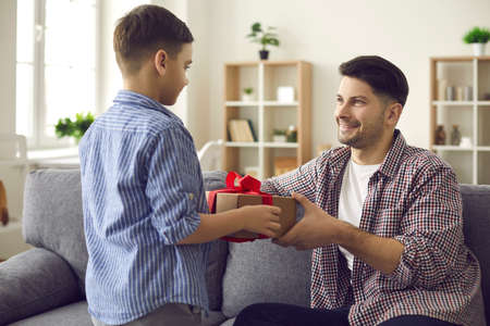 Happy smiling loving daddy sitting on sofa taking surprise gift box from adorable son hands. Birthday and fathers day holiday home celebration, parent and child love relationship and bondingの写真素材