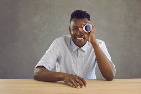 Studio portrait of happy curious black man looking with one eye through hole in paper roll. Cheerful...