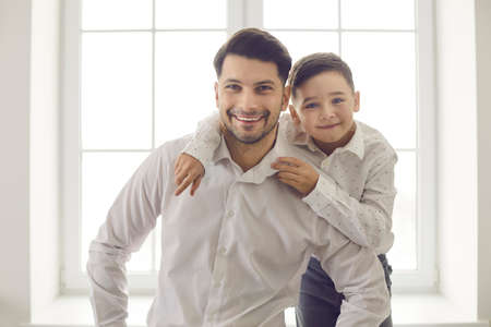 Happy fathers day. Close up portrait of cute dad with little son hugging and looking against window background. Dad spends time with his beloved son at home. Concept of family and parenthood.の写真素材
