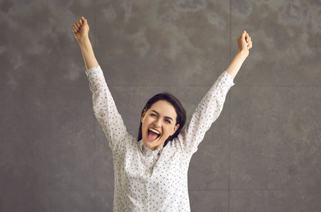 Happy cheering optimistic young casual woman screaming and rejoicing raising hand studio shot. Millennial girl having fun celebrating victory standing on grey copy space. People expressionの写真素材