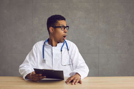 African american male doctor looks away with an astonished expression on his face. Man with a clipboard in his hands sits at a table with his mouth wide open and looks towards the free space for text.の写真素材