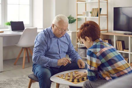 Senior concentraited man grandfather sitting and playing checkers with his grandson boy at home with room interior at background. Grandchildren spending time with grandparents conceptの写真素材