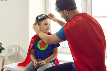 Happy family spending time together. Father and child wearing red super hero costumes having fun at home. Young dad helping smiling little son put on aviator goggles while playing fun superhero gamesの写真素材