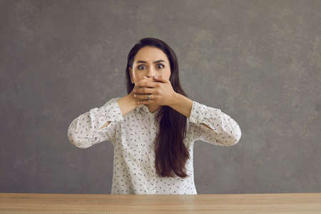 Scared young caucasian woman covering mouth with hand sitting at table studio shot. Female student feeling fear, being afraid of something showing shock and horror emotion headshot portraitの写真素材