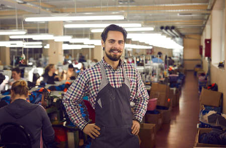 Portrait of footwear factory worker at work. Happy handsome young man in apron uniform looking at camera standing in shoe sewing workshop room. Shoe manufacturing industry conceptの写真素材