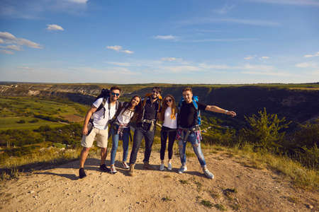 Group of young smiling travelers hikers standing with backpacks together and enjoying freedom with green valley landscape at background on sunny clear day. Hiking and traveling together.の写真素材