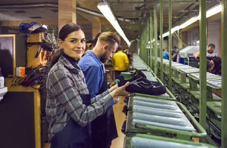 Shoe production manufactory concept. Male and female shoemaker team working at production line at footwear factory. Focus on young woman worker in uniform smiling looking at camera sorting bootsの写真素材