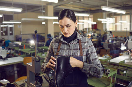 Serious female shoe factory worker checking quality of footwear. Woman holding black mens leather boot, looking at seams and checking stitches. Shoe manufacturing industry conceptの写真素材
