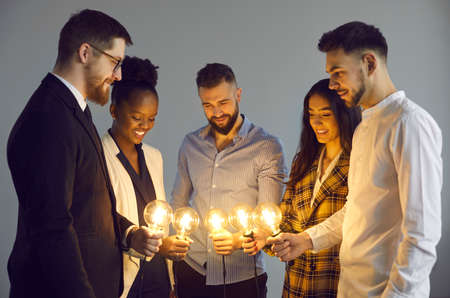 Group of happy mixed-race colleagues join Edison light bulbs as symbol of sharing ideas. Team of young business people holding shining lightbulbs as metaphor for creative community, teamwork and unityの写真素材