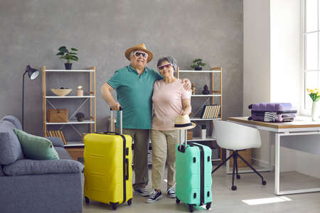 Portrait of happy senior tourists with packed suitcases standing in living room at home. Active retired elderly husband and wife ready for summer vacation. Traveling and tourism conceptの写真素材