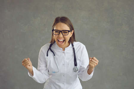 Happy excited overjoyed woman in white nurse or doctors lab coat shouting Yes celebrating success, victory, job placement, salary pay rise, finding cure for patient, or end of Covid 19 virus pandemicの写真素材
