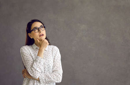 Headshot portrait young caucasian woman thinking touching chin with hand standing isolated on grey background. Girl student or female freelance employee planning or dreaming about futureの写真素材