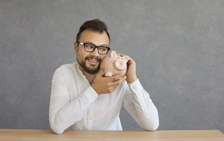 Happy excited smiling adult caucasian business man sitting at table holding and hugging piggybank against grey background. Saving money, finance investment, budget planning and saving conceptの写真素材