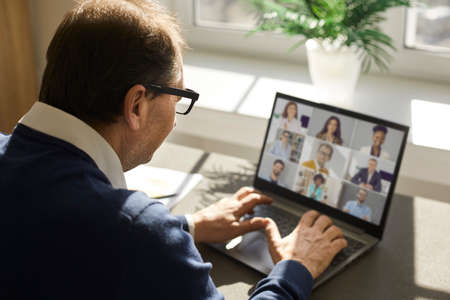 Senior man sitting at desk, looking at laptop computer and having virtual video call group meeting with team of employees and coworkers. Online communication, teamwork and business management conceptsの写真素材