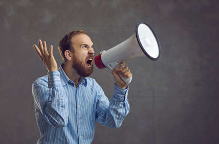 Agitated young man in blue shirt shouting loudly in megaphone. Excited salesperson, office worker or...