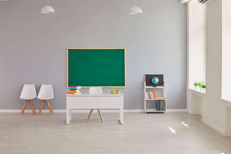 Interior of modern light empty spacious school classroom with clean green blackboard, teachers desk, chairs, shelf, books and earth globe. Education, back to school conceptの写真素材