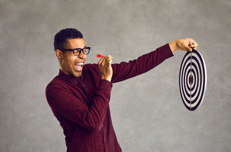 Side view smiling confident young black man aiming red dart at shooting target determined to throw it and hit bullseye. Using chance, accepting challenge, achieving business goal and objective conceptの写真素材