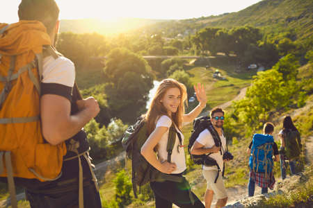 Group of happy backpackers trekking on sunny day. Young tourists traveling and enjoying active summer vacation. Smiling woman looking at camera and waving hand walking down hiking trail with friendsの写真素材