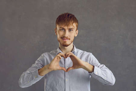 Caucasian red-haired man making heart gesture showing love and charity studio portrait shot. Happy smiling romantic young casual guy expressing affection and admiration. Positive people emotionの写真素材