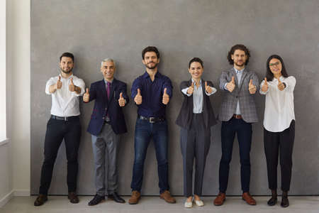 Happy business leaders or group of employees showing thumbs up while standing against gray wall and looking at camera. Professional office team recommends the best corporate service or a good career.の写真素材