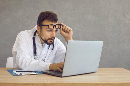 Male doctor with a shocked and confused expression looks at a laptop screen seeing a medical error. Man with frightened expression looks out from under glasses sitting at a table on a gray backgroundの写真素材