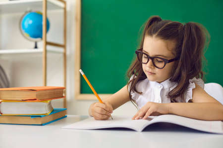 Back to school concept. A smart girl in glasses writes with a pencil in a notebook while sitting at...