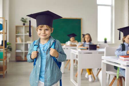 Portrait of happy school child. Successful elementary school graduate in academic grad hat standing in classroom. Cute little boy in mortarboard looking at camera and smilingの写真素材