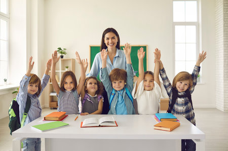 Portrait of cheerful female teacher and little students with bags standing together near table in modern classroom. Happy school kids and their tutor smiling, raising hands and looking at cameraの写真素材