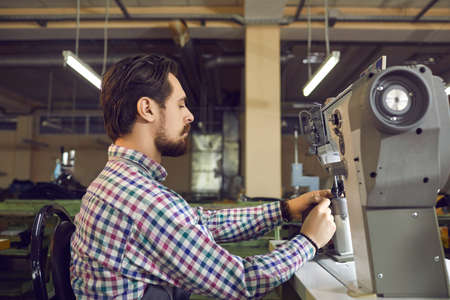 Male worker making details for new boots. Profile portrait of a serious young man working on an industrial sewing machine at the workshop of a shoe factory. Footwear manufacturing industry conceptの写真素材