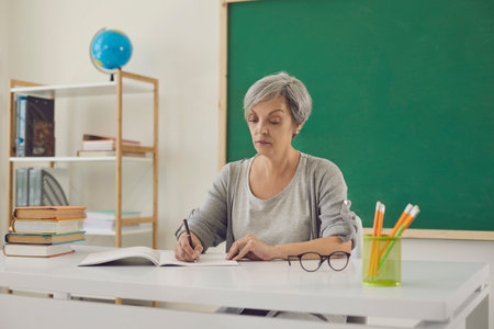 Woman teacher writes while sitting at desk in classroom with green board at school. Education...