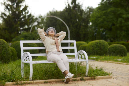 Happy relaxed senior lady sitting on wooden bench looking in sky enjoy sunny day in public city park. Carefree emotion and daydreaming, healthy lifestyle and rest on retirement conceptの写真素材