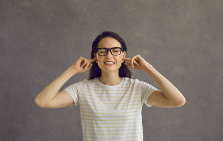 I cant hear you. Head shot of young woman in glasses closing eyes and plugging ears with fingers to ignore loud noise, unwanted opinion, dumb stupid comment or useless unsolicited uninvited criticismの写真素材