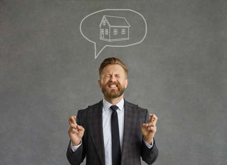 Positive emotions. Successful man with a funny expression crosses his fingers dreaming of his own house. Middle-aged Caucasian man stands on a gray concrete background and hopes for the best miracleの写真素材