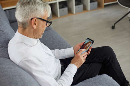 Concept of older adults using online dating apps or websites. Old bachelor searching for future wife. Senior man holding mobile phone in hand and looking at photo of beautiful young woman on displayの写真素材