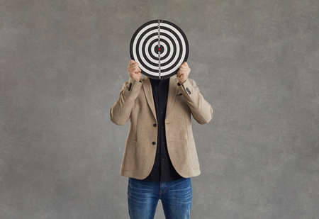 Unknown businessman hid his face behind a darts board while standing on a gray background. Concept of setting business goals, finding the target audience, achieving goals and successの写真素材
