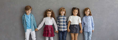 Banner with portrait of happy little kids in casual wear. Group of adorable children in new neat clothes holding hands, smiling and looking at camera standing together against grey studio backgroundの写真素材