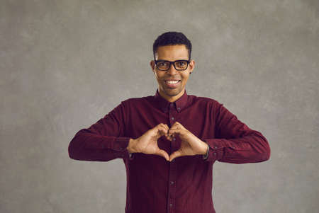 Portrait of young smiling african american guy making heart gesture while standing on gray background. Friendly man expresses his love. Concept of romance, body language and declaration of love.の写真素材