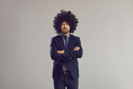 Serious adult caucasian bearded hipster business man in stylish curly hair wig and formal suit standing with arms crossed looking at camera studio portrait shot isolated on grey background.の写真素材