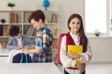 Back to school, kids learning in group. Caucasian elementary schoolgirl in uniform standing with backpack and copy-book in hand looking at camera lifestyle portrait shot. Classroom and classmatesの写真素材